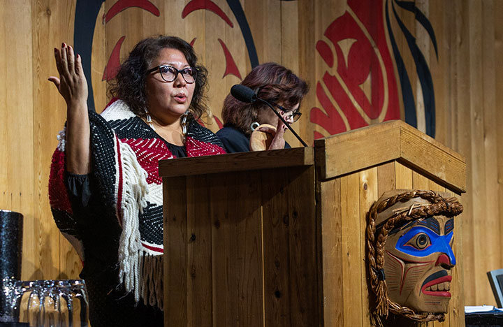 Panelists on stage speaking at a lectern with a carved mask on the front